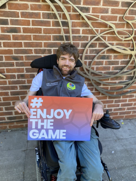 Person sitting in power wheelchair holding a sign that reads Enjoy The Game